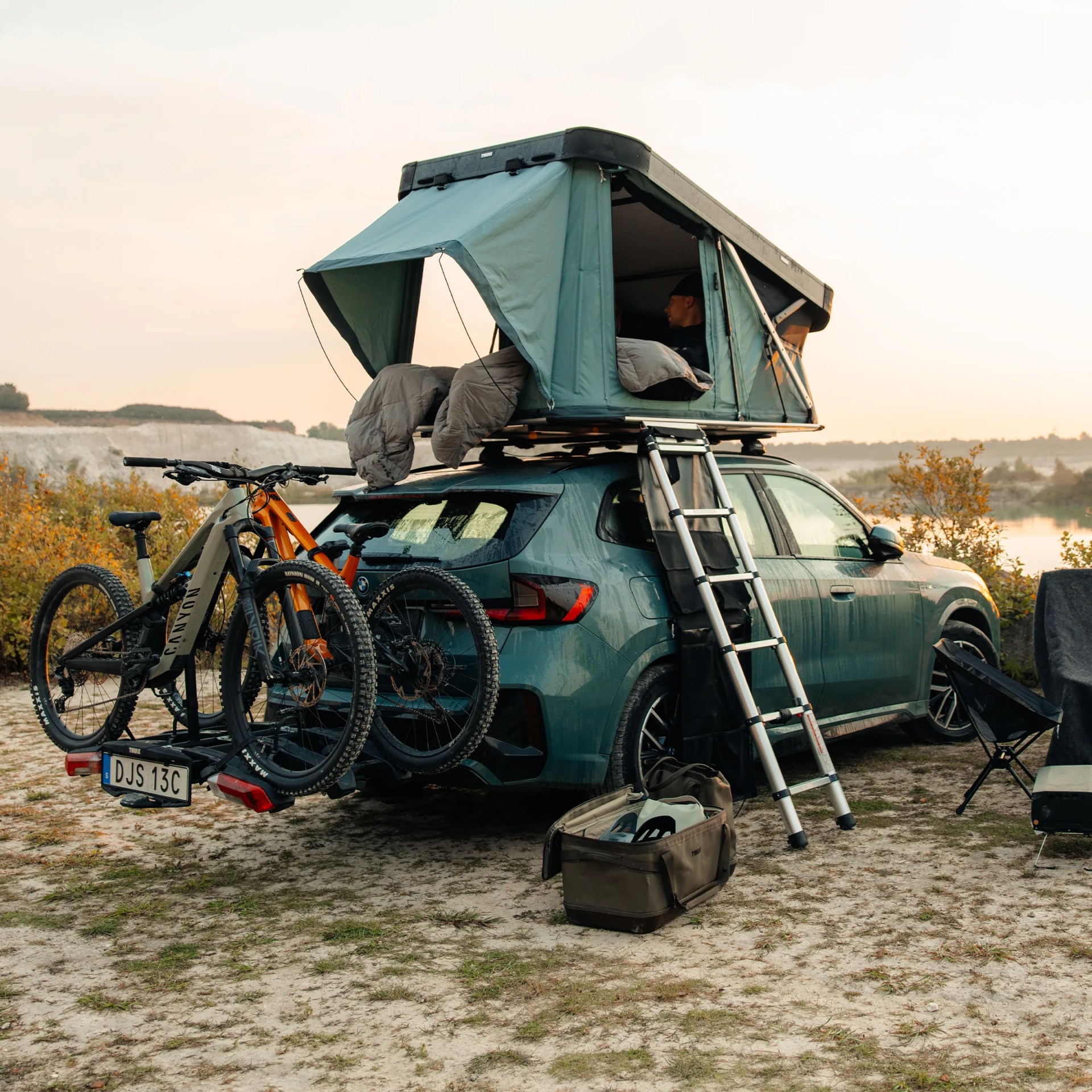 Green SUV with rooftop tent and ladder, two mountain bikes mounted on rear rack, and camping gear nearby.