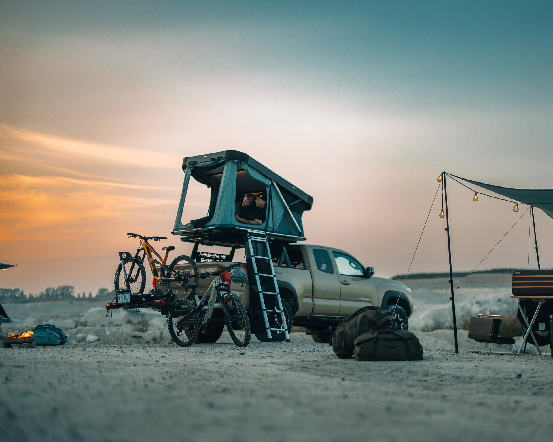 Beige pickup truck with rooftop tent and two mountain bikes parked beside it at sunset campsite.