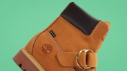 Tan leather boot with black padded collar, white stitching, gold buckle, and embossed tree logo on a green background.