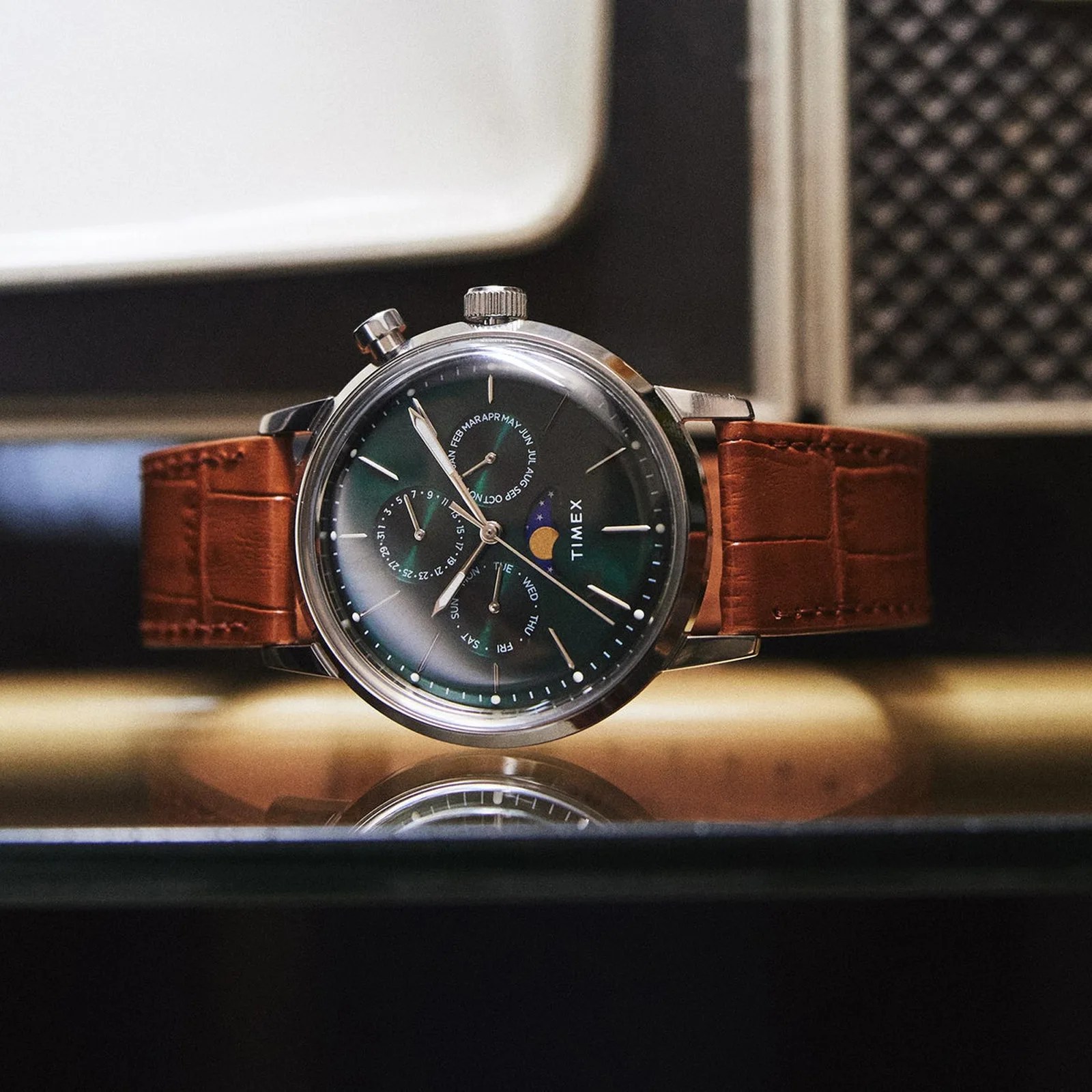 Timex wristwatch with a green dial, moon phase indicator, and brown leather strap on a reflective surface.