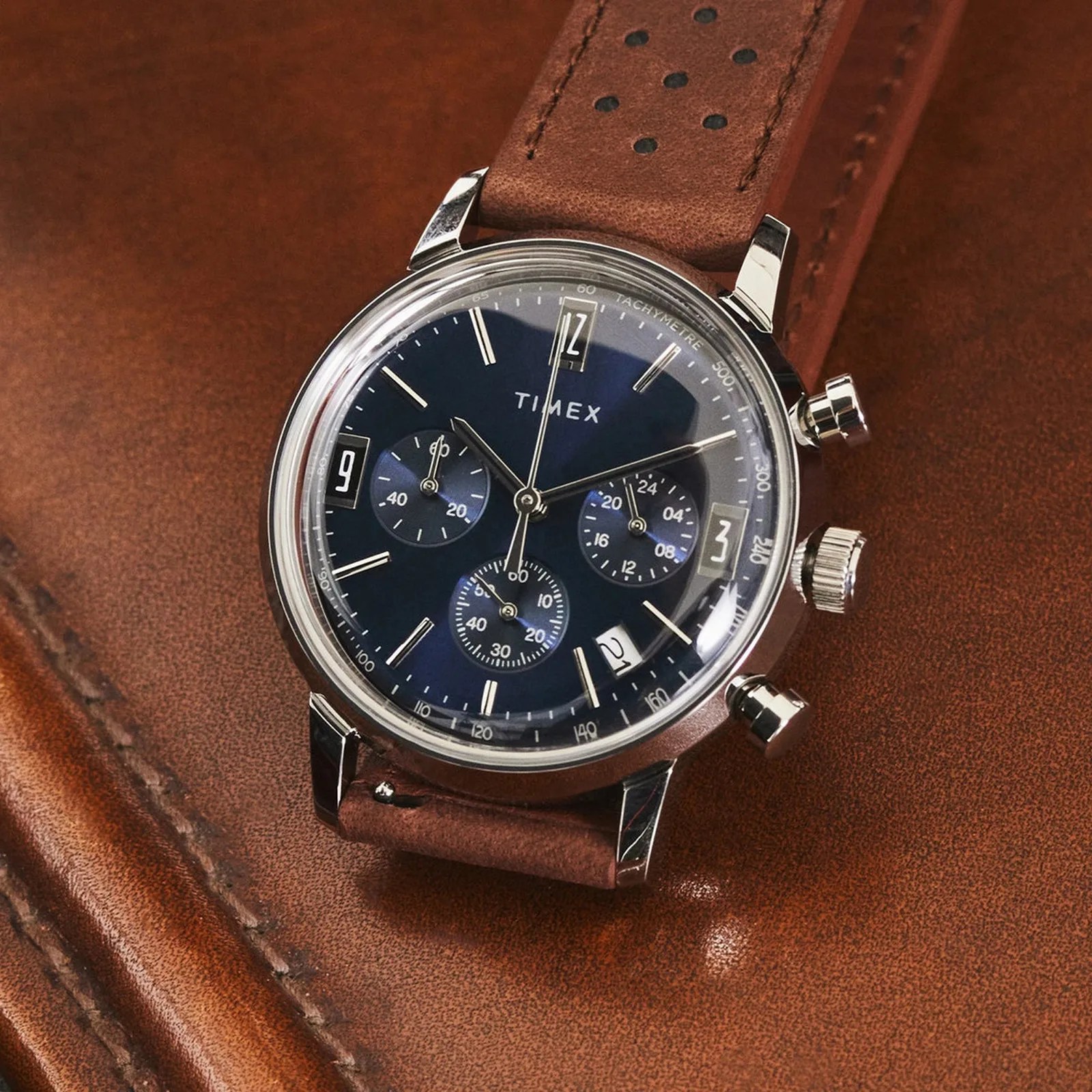 Timex chronograph watch with blue dial, silver case, and brown perforated leather strap on brown leather surface.