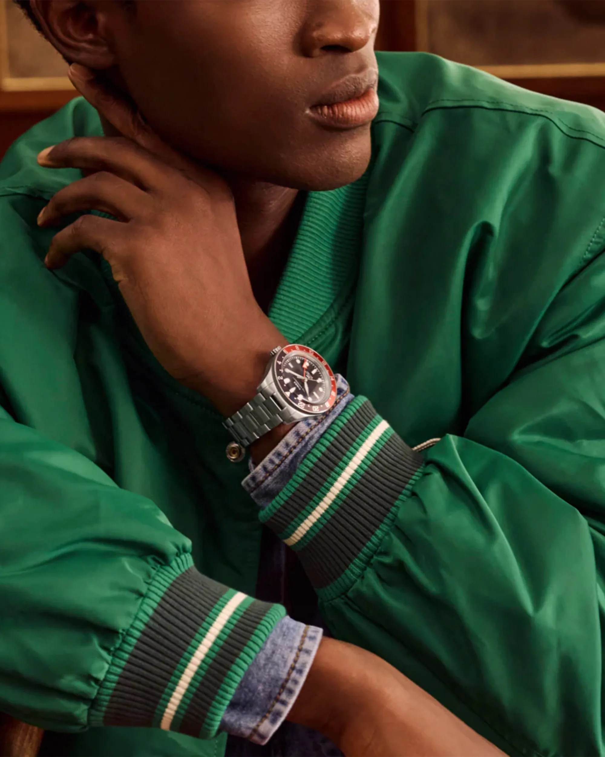 Silver metal wristwatch with black and red bezel worn on wrist over denim shirt and green jacket.