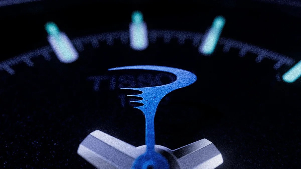 Close-up of a blue luminescent watch hand on a black Tissot watch face with glowing hour markers.
