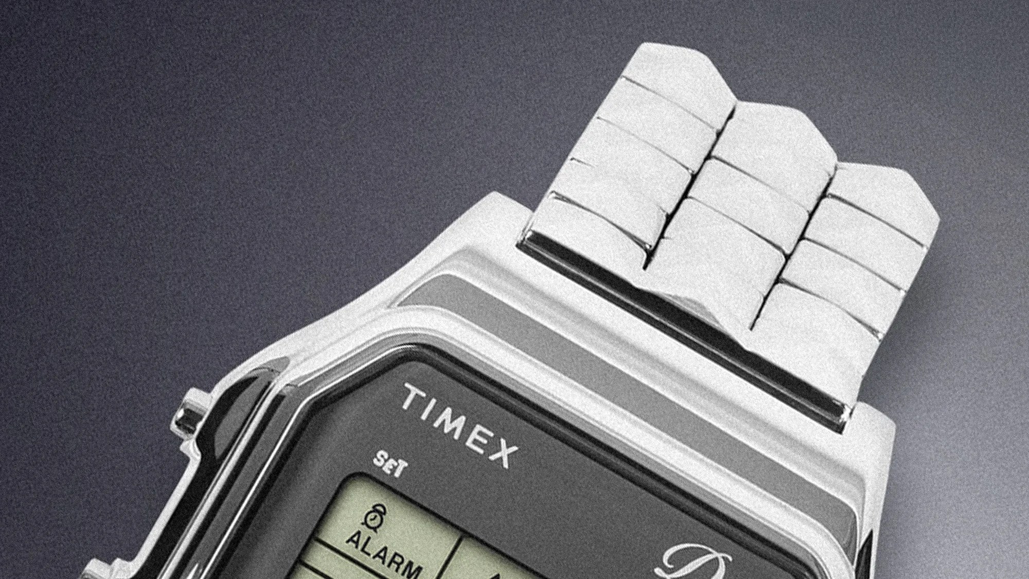 Close-up of a silver Timex digital watch with a metal link band on a gray background.