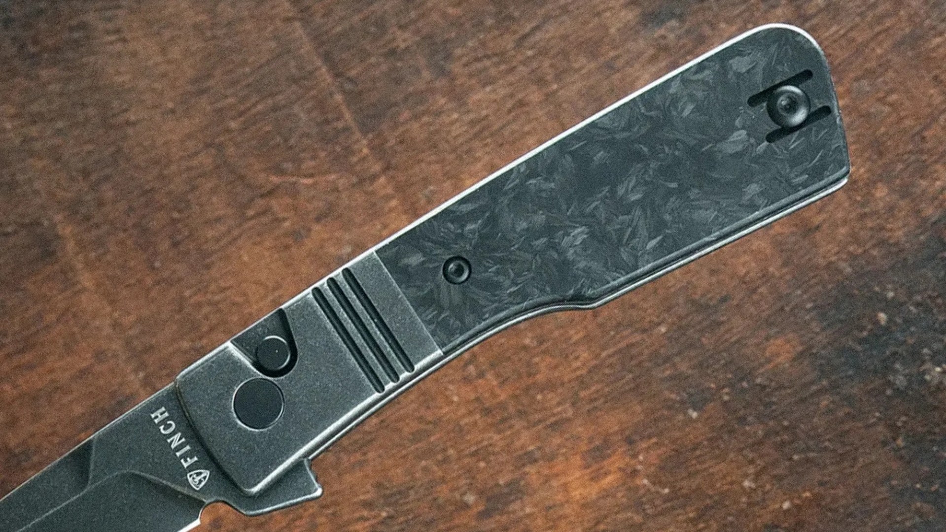 Close-up of a black folding knife handle with textured grip and "FINCH" logo on a wooden surface.