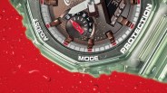 Close-up of a translucent green Casio G-Shock watch face with brown and red accents, showing day indicator and mode buttons, on a red surface with water droplets.