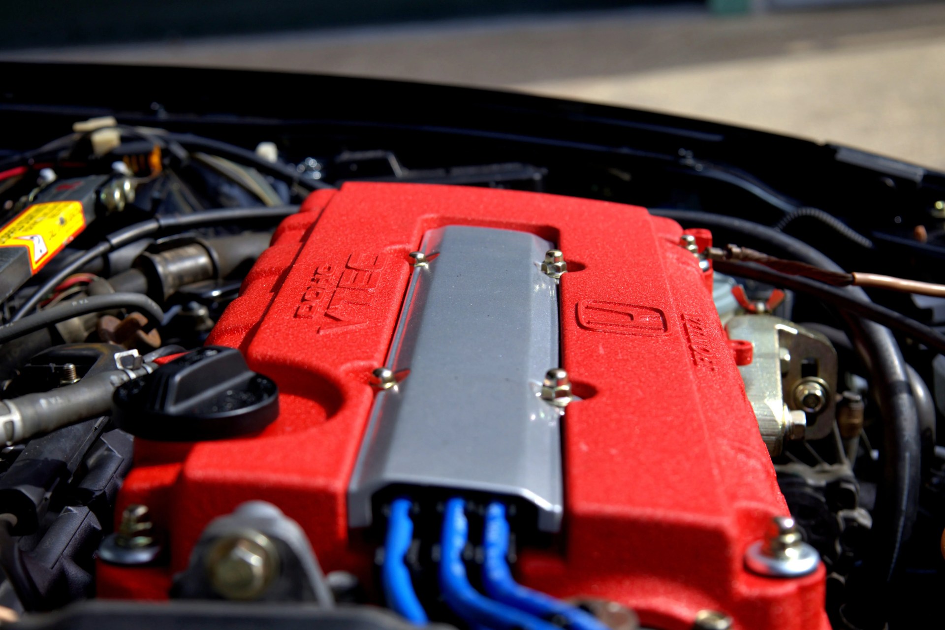 Red DOHC VTEC engine cover with silver center and blue spark plug wires in a car engine bay.