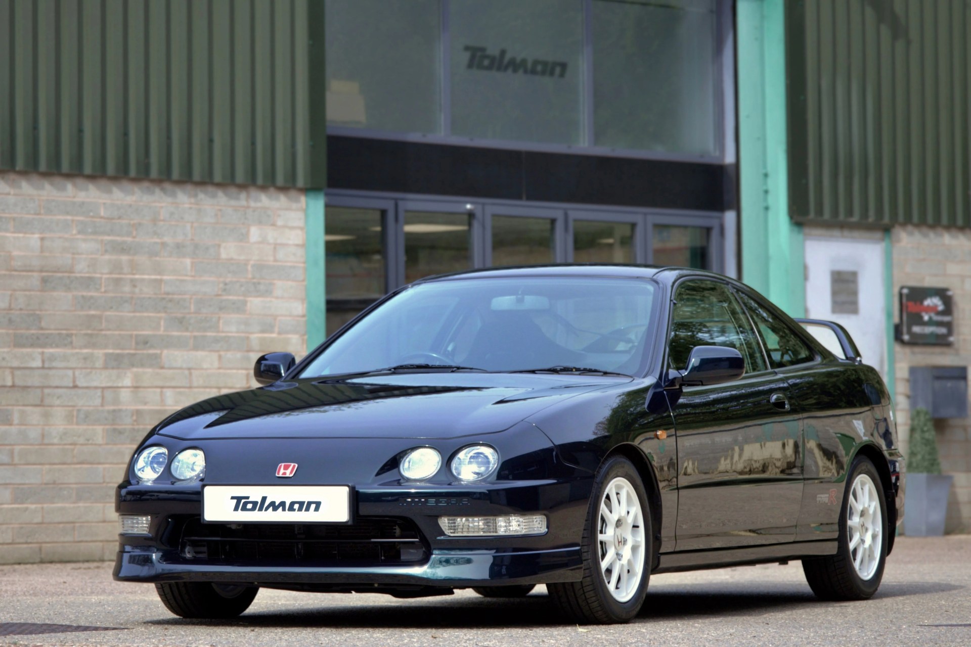 Black Honda Integra Type R coupe with white wheels parked in front of a building with "Tolman" signage.
