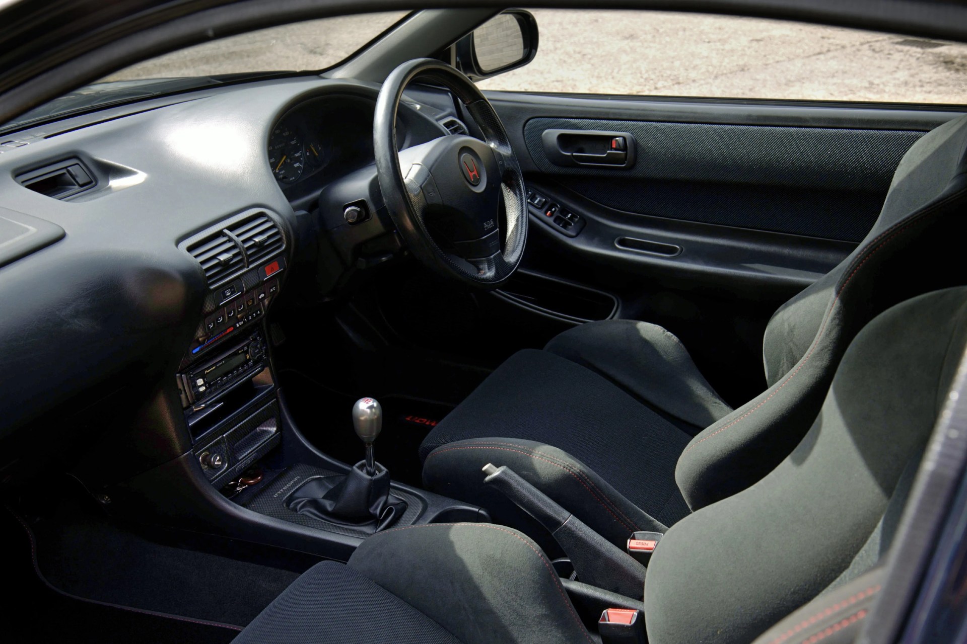 Black and gray car interior with manual gear shift, racing seats, and a steering wheel featuring a red Honda logo.