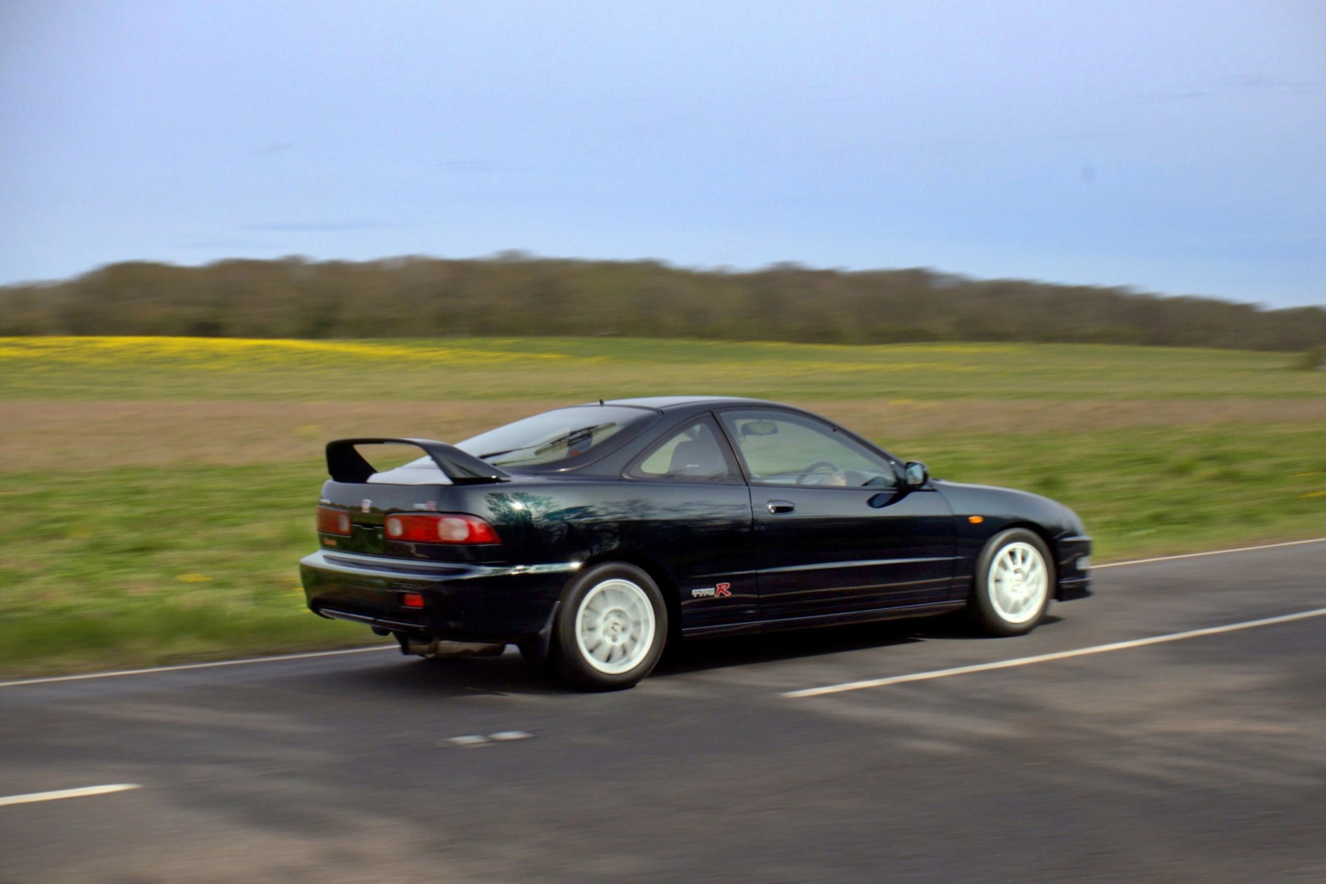 Black Honda Integra Type R coupe with white wheels driving on a rural road.