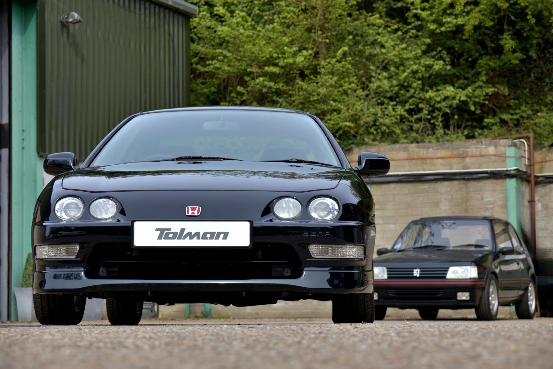 Black Honda Integra with Tolman license plate in front of a black Peugeot hatchback near a green building and brick wall.