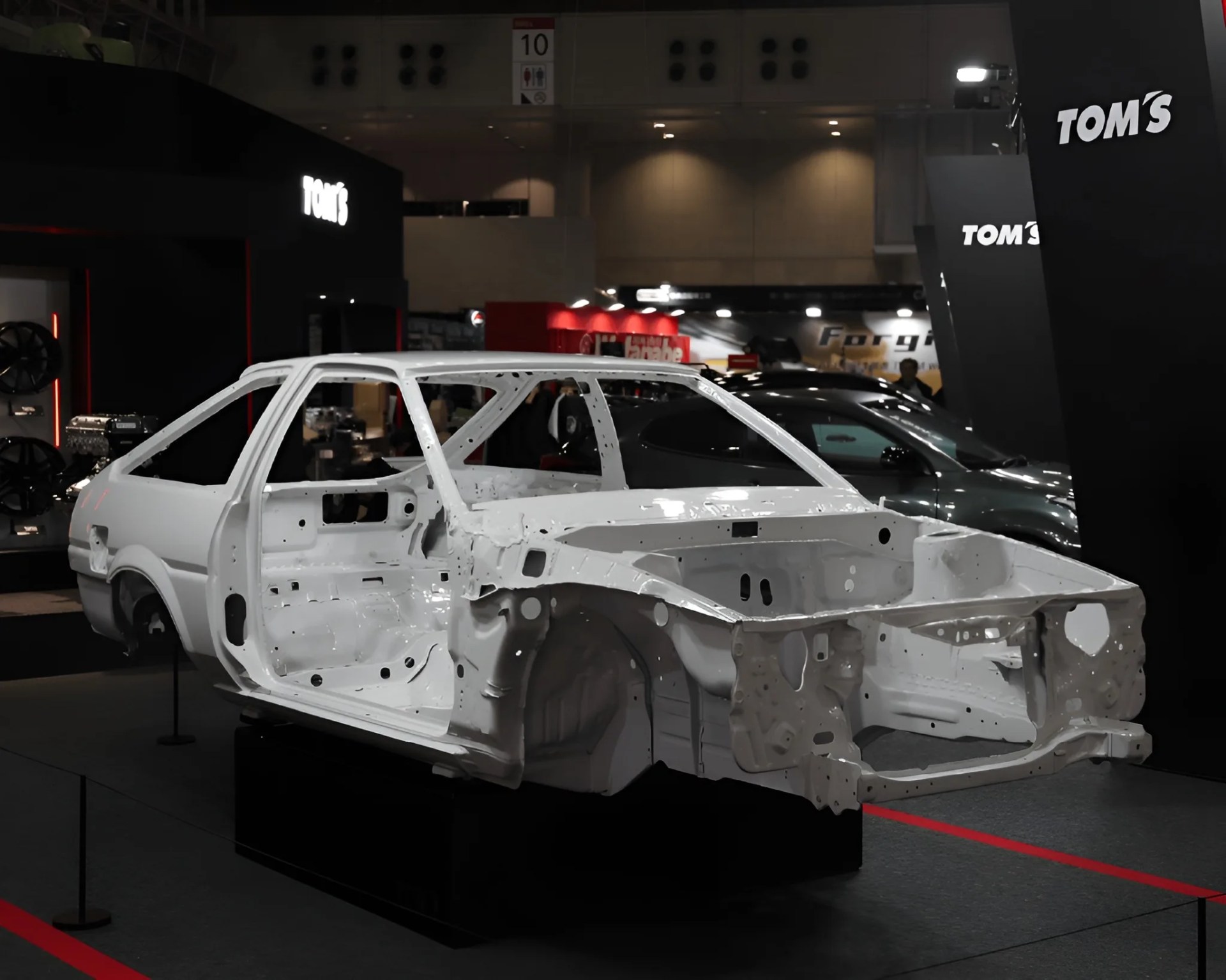 White bare car chassis without doors or engine displayed indoors with "TOM'S" branding in the background.
