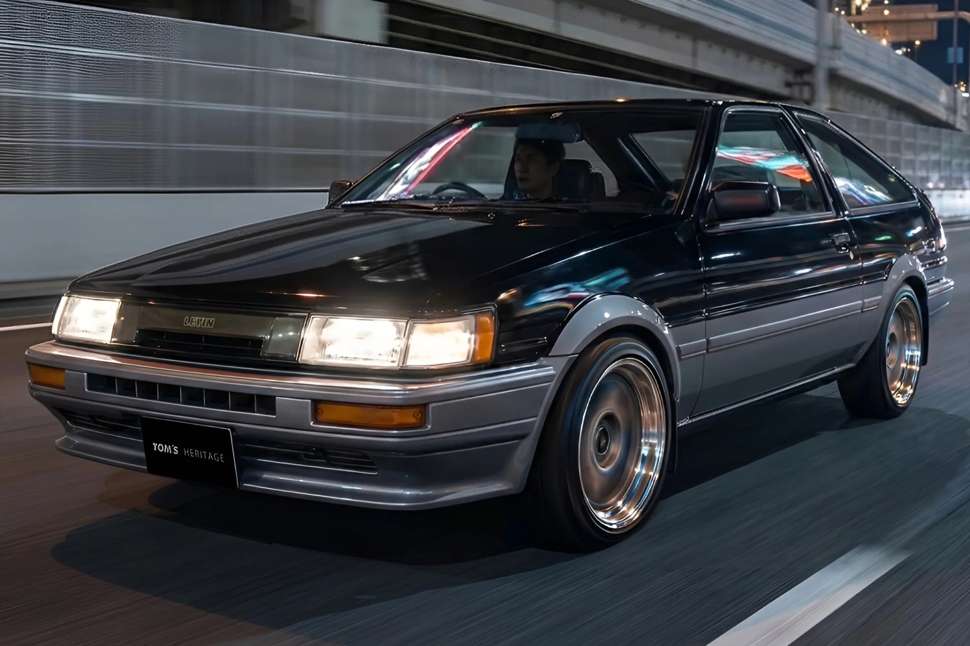 Black and gray two-tone Toyota Levin hatchback with headlights on driving on a city street at night.