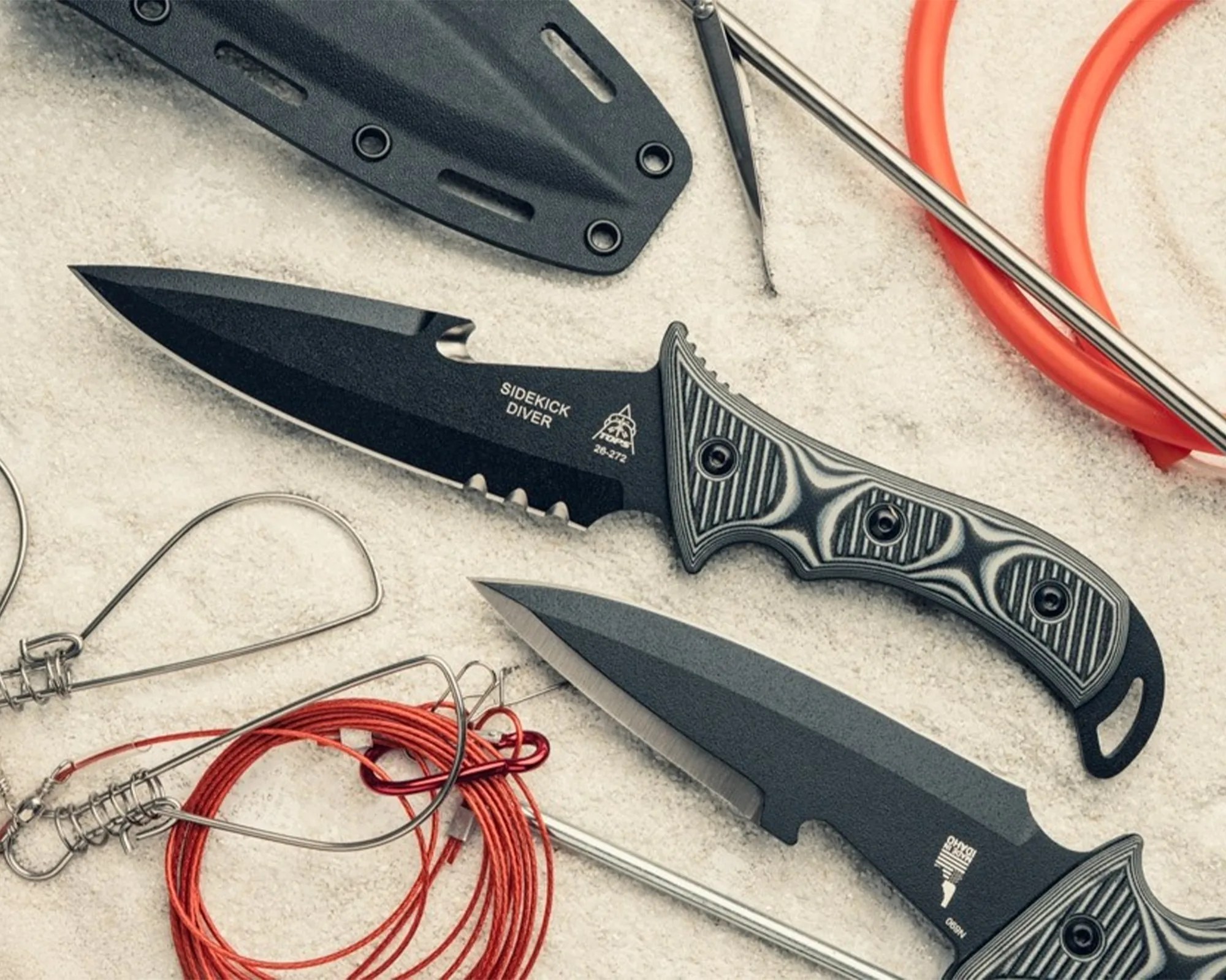 Two black diving knives with textured handles and serrated edges lying on sand next to red fishing lines and metal fishing hooks.