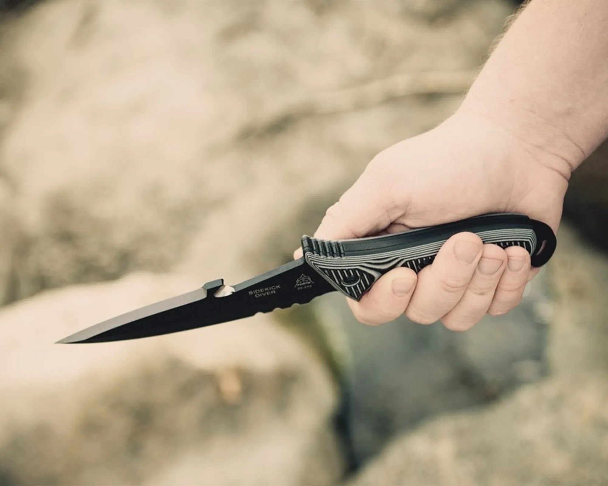 Hand holding a black tactical knife with a textured grip and partially serrated blade.
