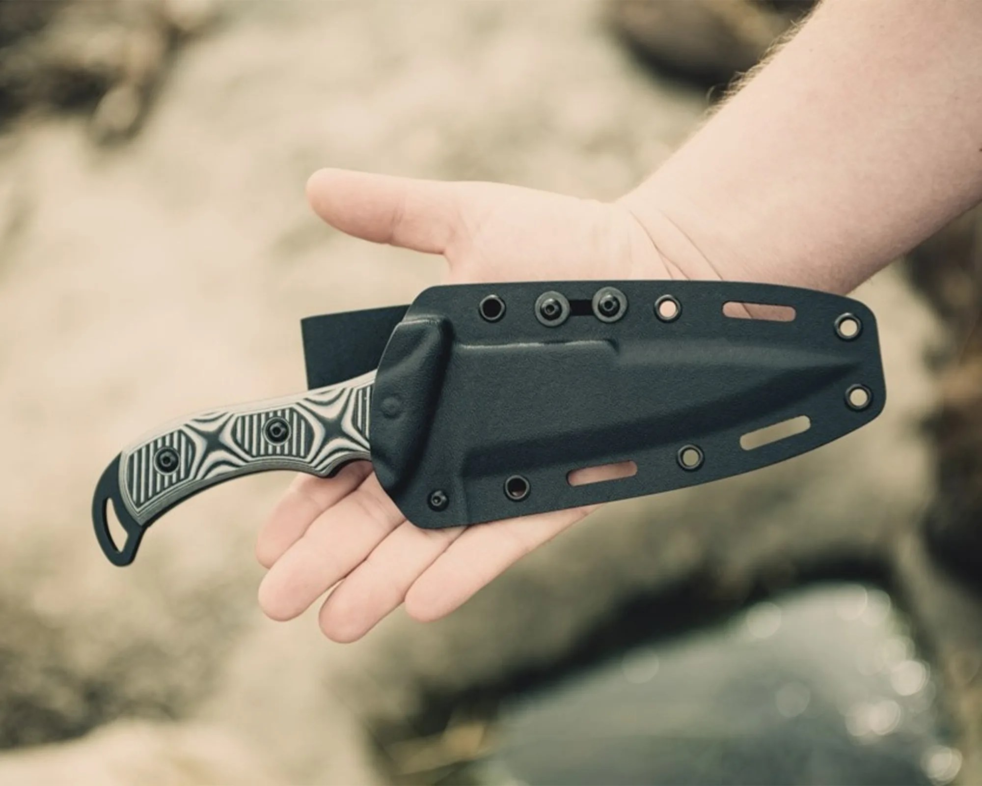Hand holding a knife with a black sheath and a patterned handle.