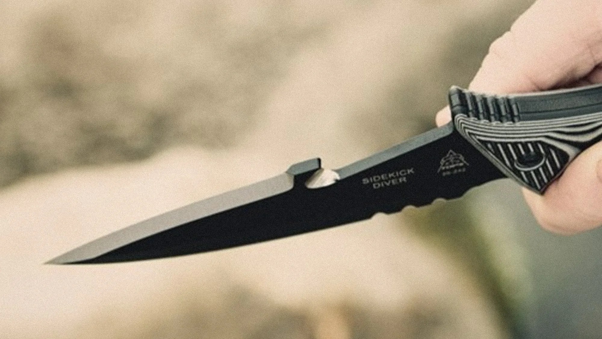 Black tactical knife with serrated edge and patterned handle held in a hand.