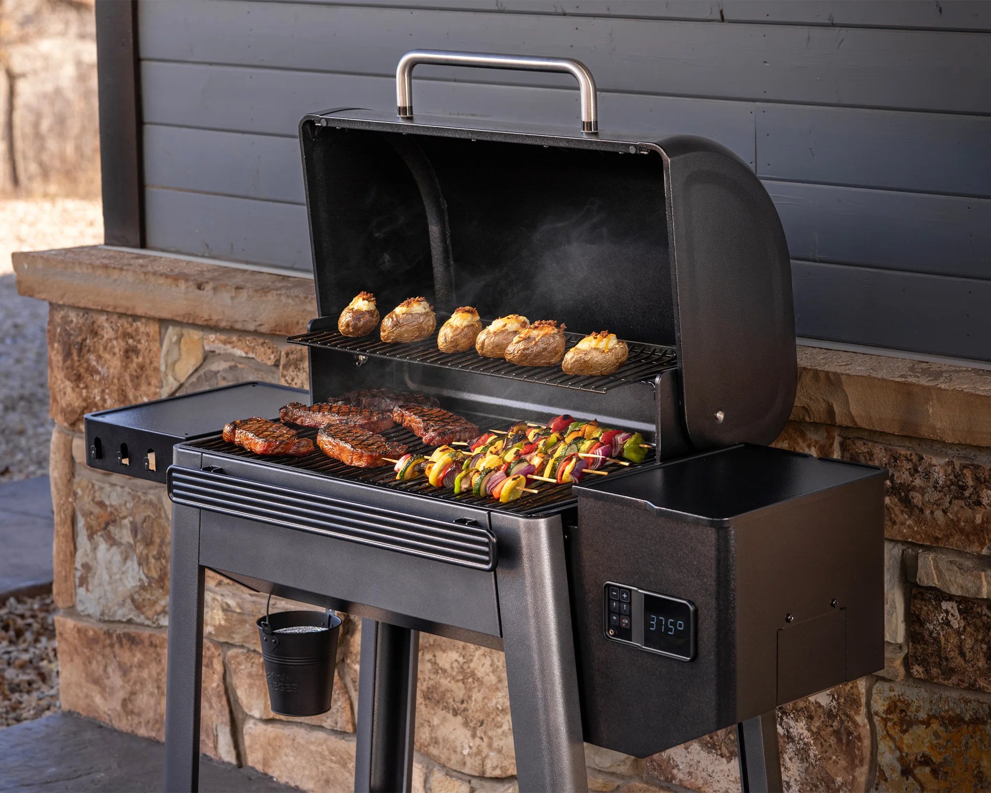 Black pellet grill with open lid showing baked potatoes, steaks, and vegetable skewers cooking, digital temperature display reading 375°F.