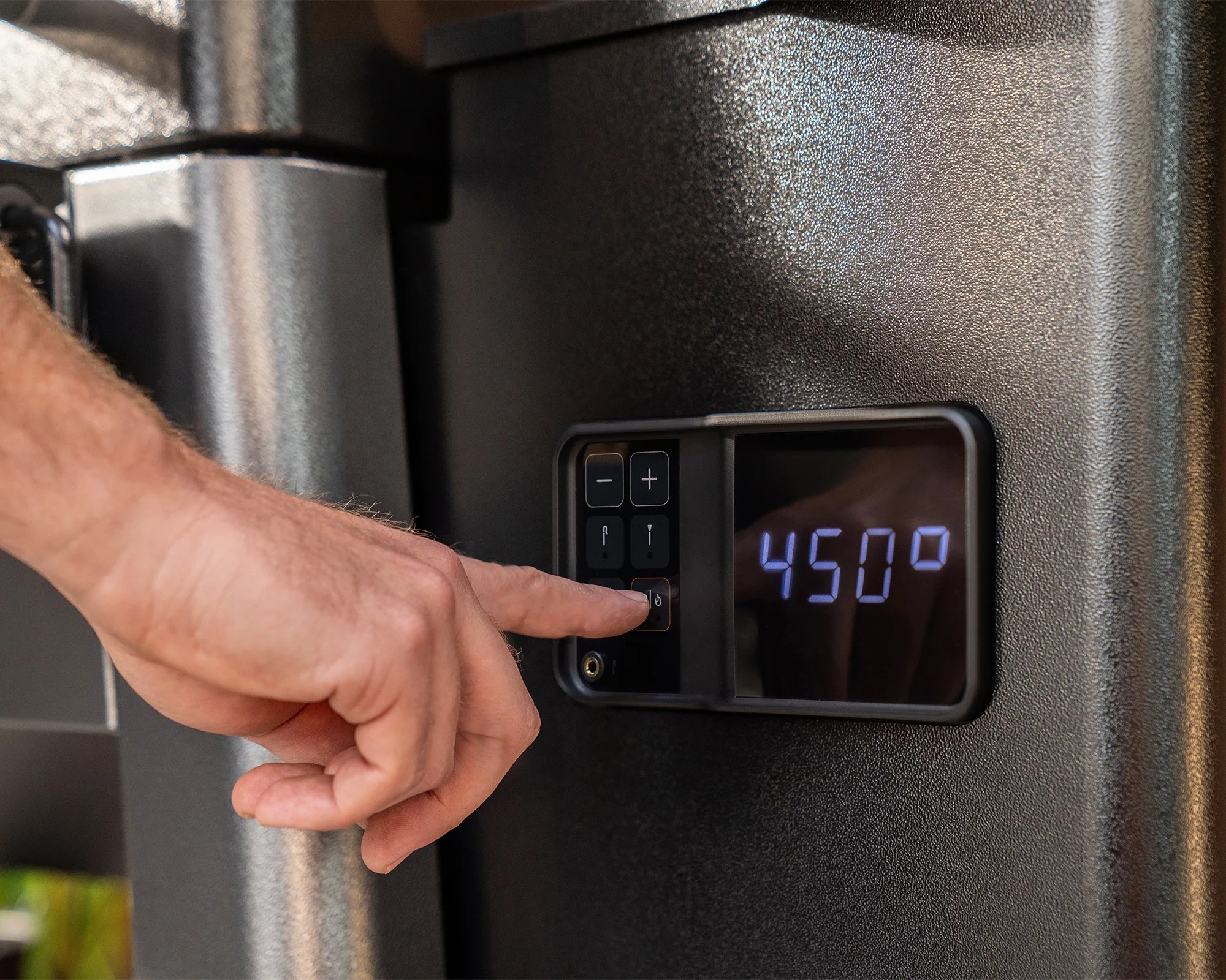 Hand pressing power button on a black digital control panel displaying 450 degrees on a textured black surface.