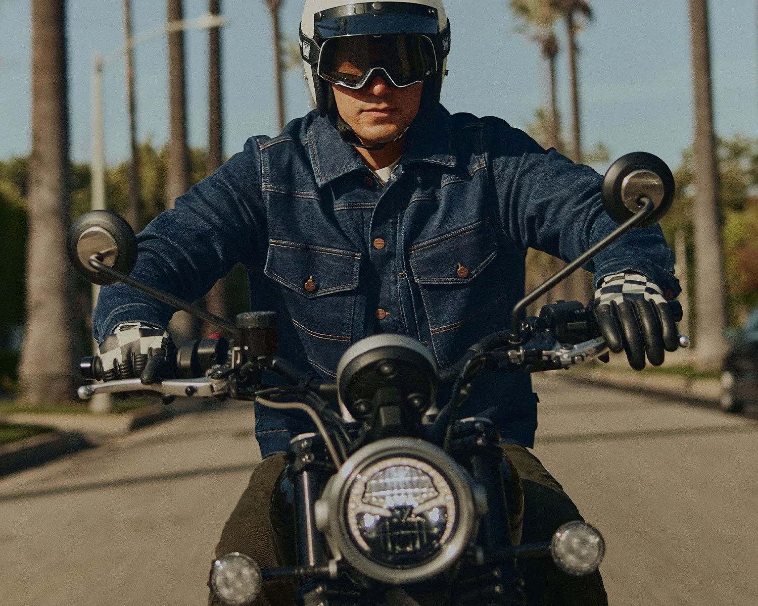Person wearing a white helmet, black goggles, denim jacket, and checkered gloves riding a motorcycle on a street.