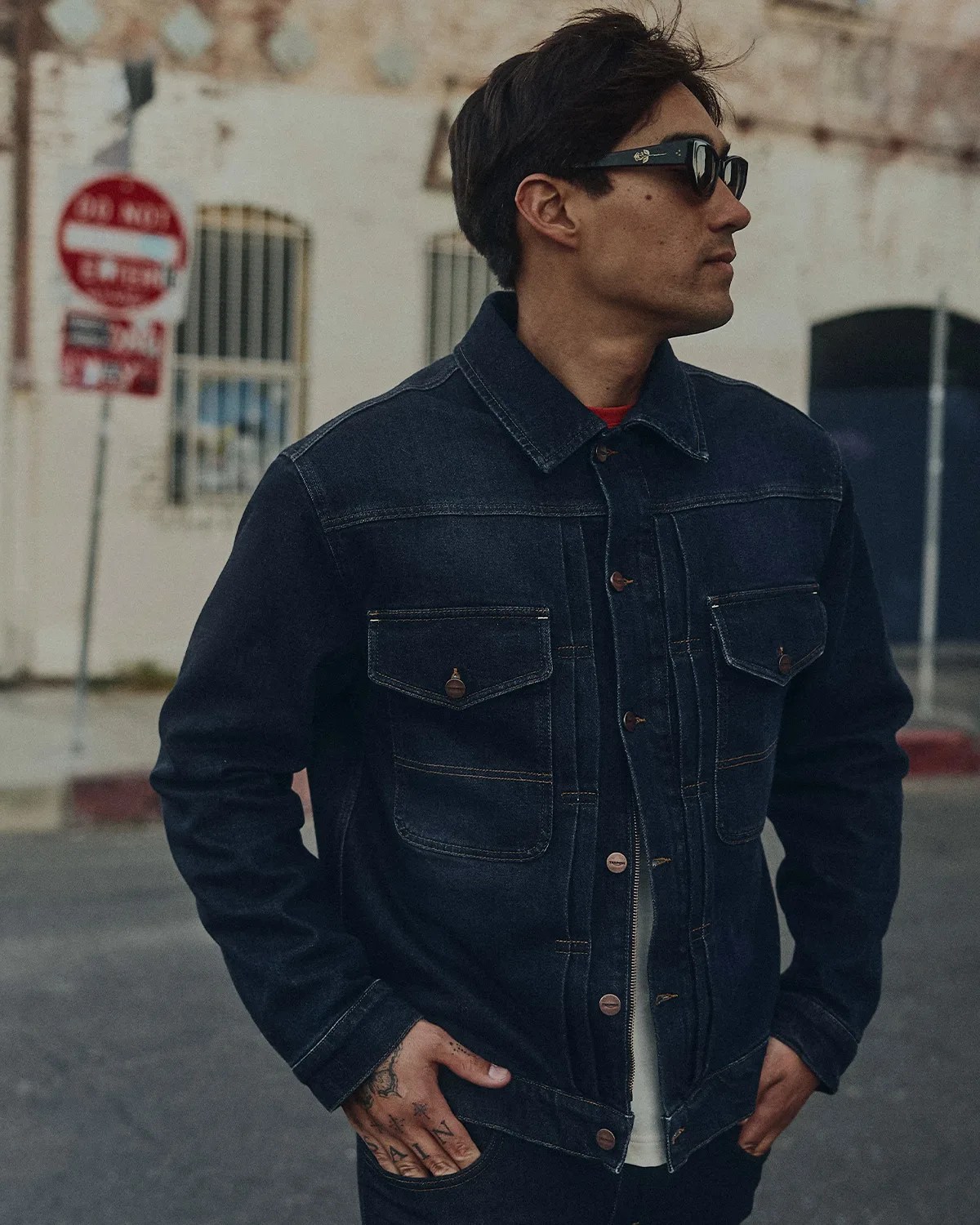 Man wearing a dark denim jacket with buttoned chest pockets and black sunglasses.