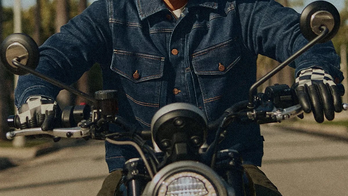 Person wearing a dark denim jacket and gloves gripping the handlebars of a motorcycle.