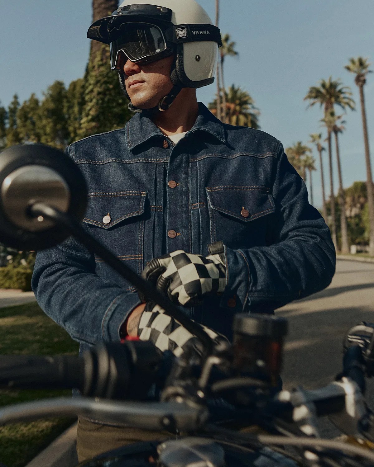 Man wearing a white helmet, black goggles, denim jacket, and checkered gloves sitting on a motorcycle with palm trees in the background.