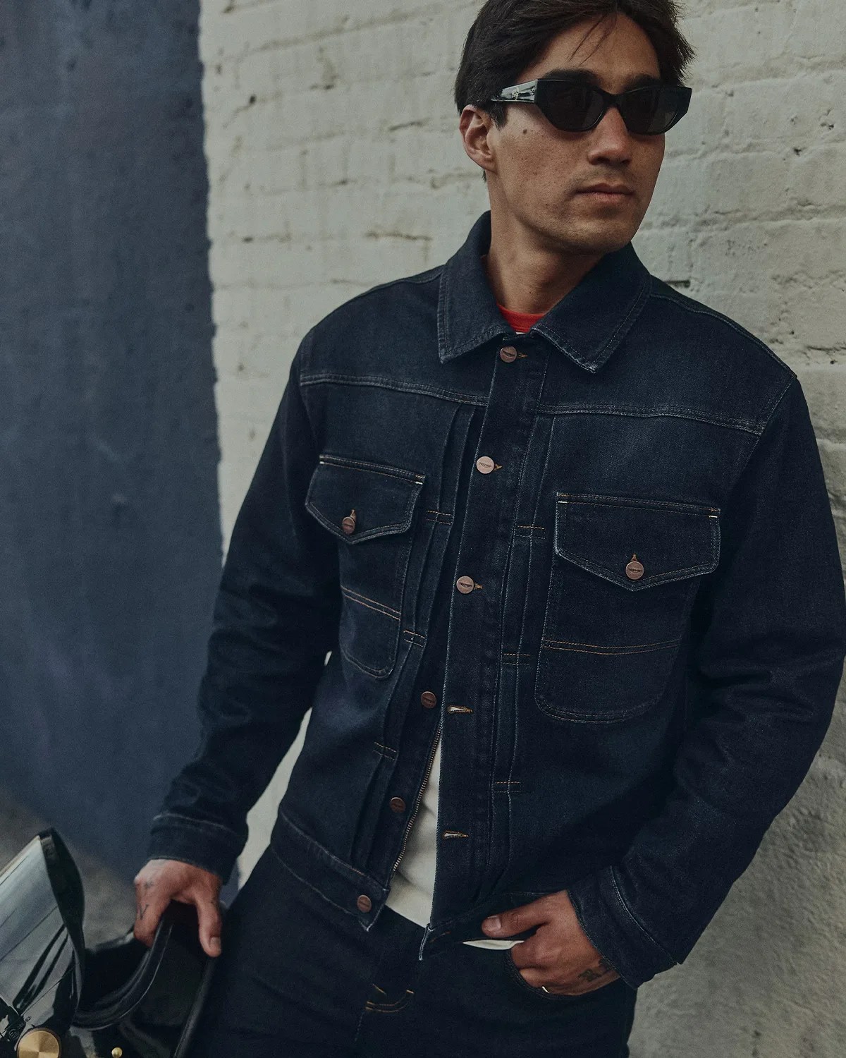Man wearing a dark denim jacket with buttoned chest pockets and black sunglasses, standing by a white brick wall.