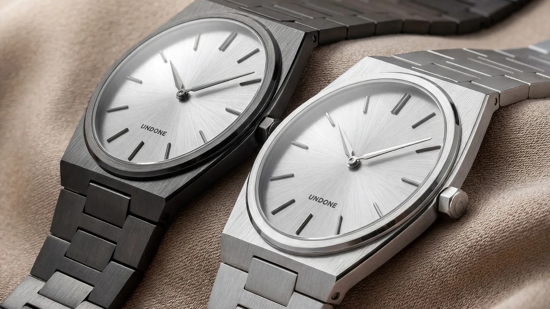 Two minimalist stainless steel wristwatches with silver dials and metal link bracelets on a beige fabric.