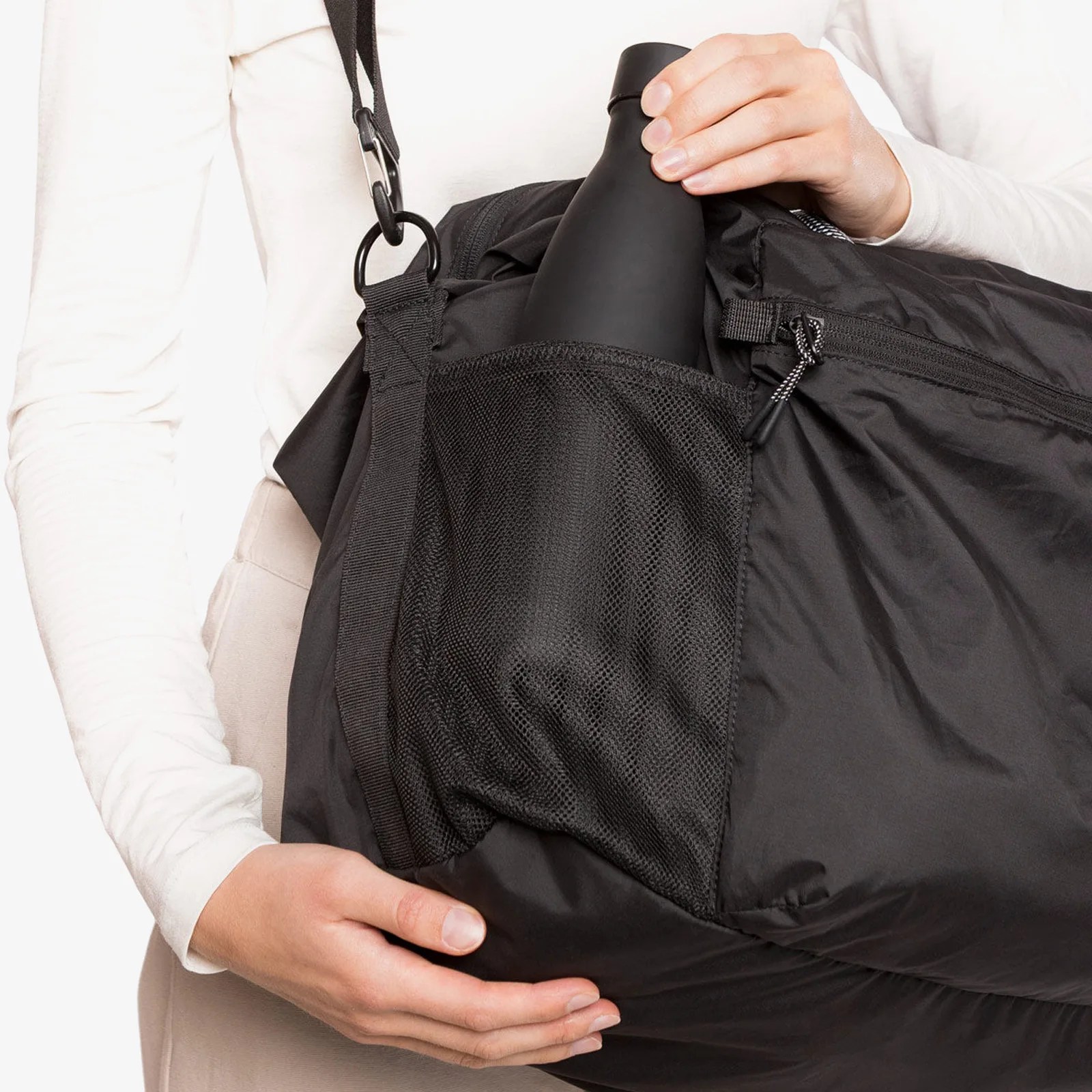 Black insulated water bottle being placed into the mesh side pocket of a black duffel bag worn by a person in a white long-sleeve shirt.