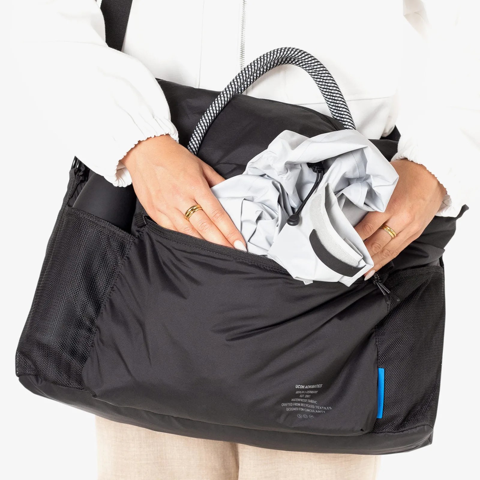 Black Ucon Acrobatics bag with mesh side pockets and a blue tag, held by a person wearing a white jacket.