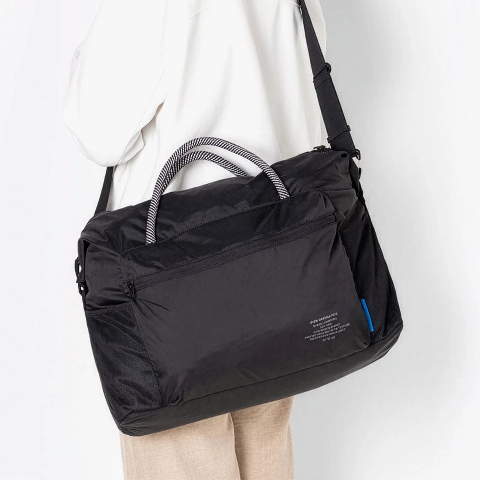 Black nylon shoulder bag with striped handles and front zipper pocket worn by a person in a white top and beige pants.