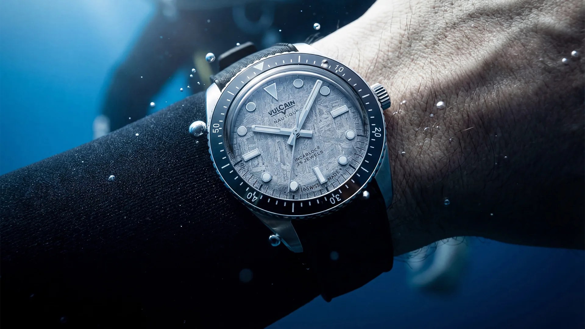 Silver dive watch with textured gray dial and black bezel worn underwater on a wrist with a black strap.