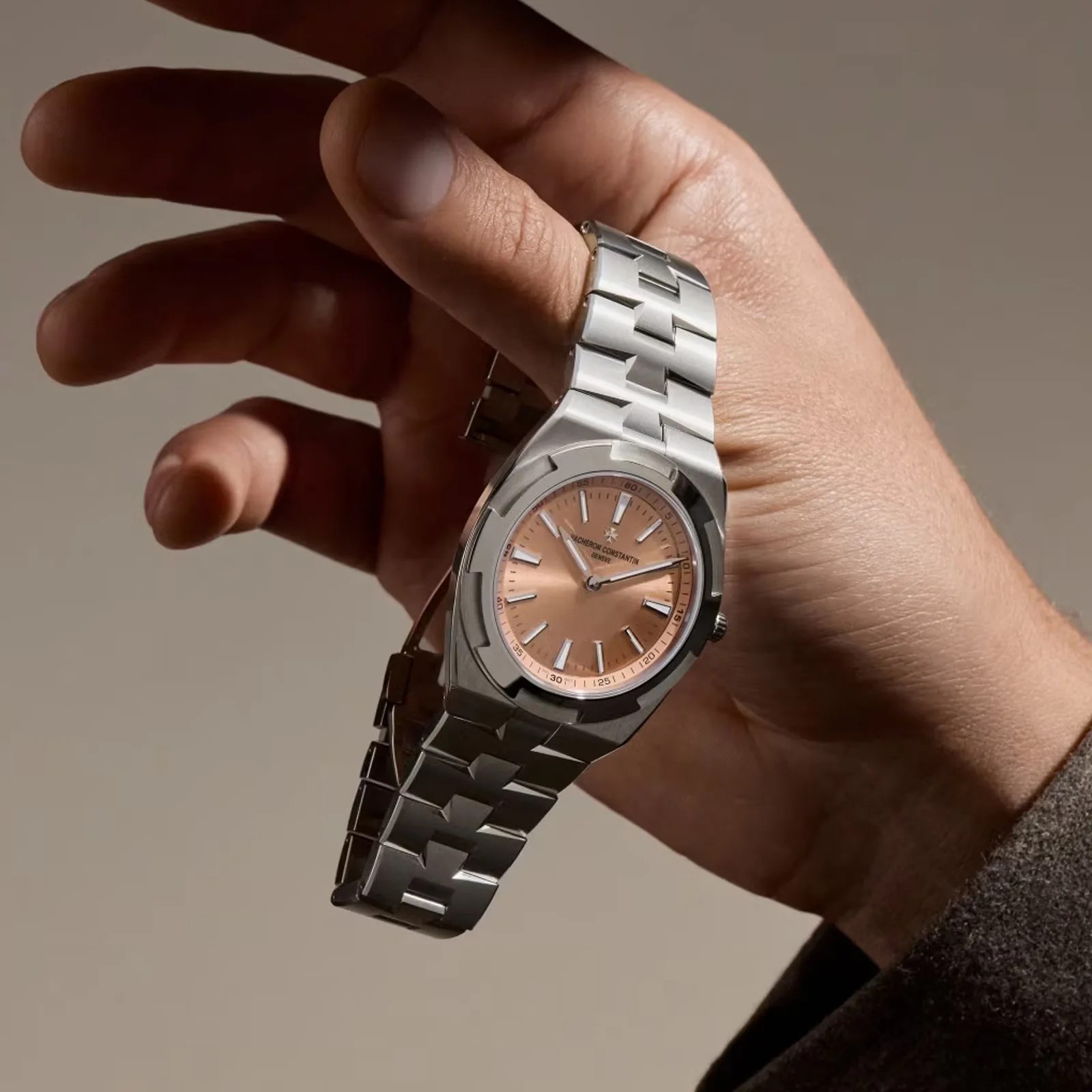 Silver metal wristwatch with a salmon-colored dial held in a hand against a neutral background.