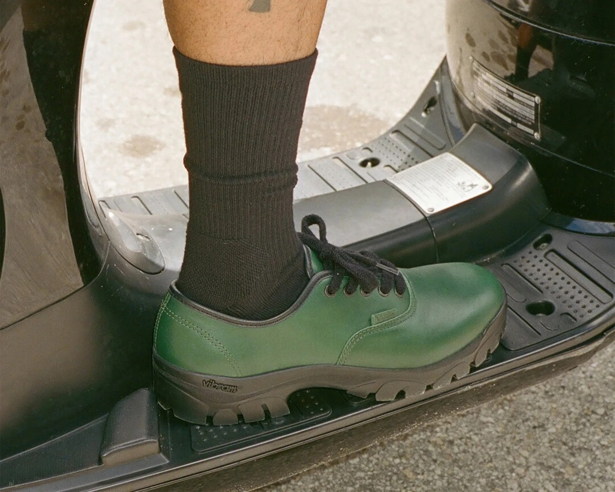 Green leather shoe with black laces and Vibram sole worn with black socks on a scooter footrest.