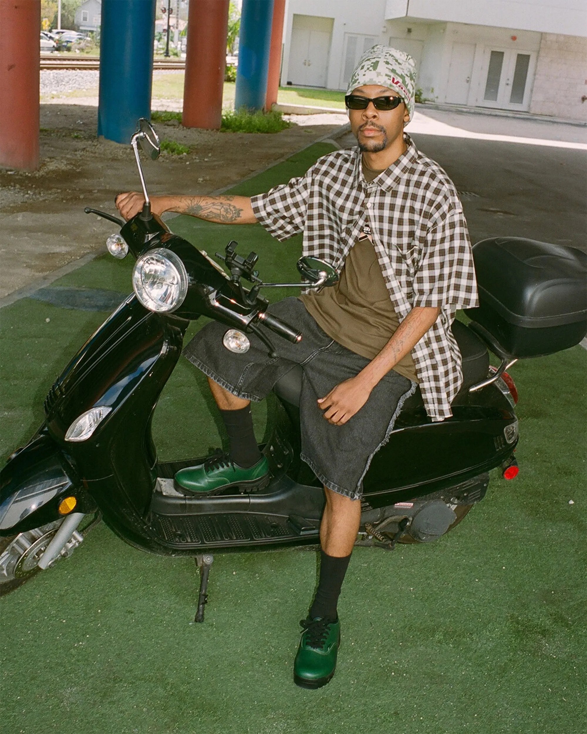 Person wearing a checkered shirt, green shoes, and a beanie sitting on a black scooter on green turf.
