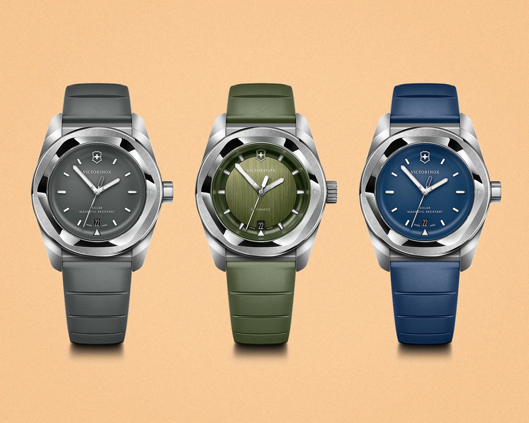 Three Victorinox wristwatches with stainless steel cases and rubber straps in gray, green, and blue, displayed on a beige background.