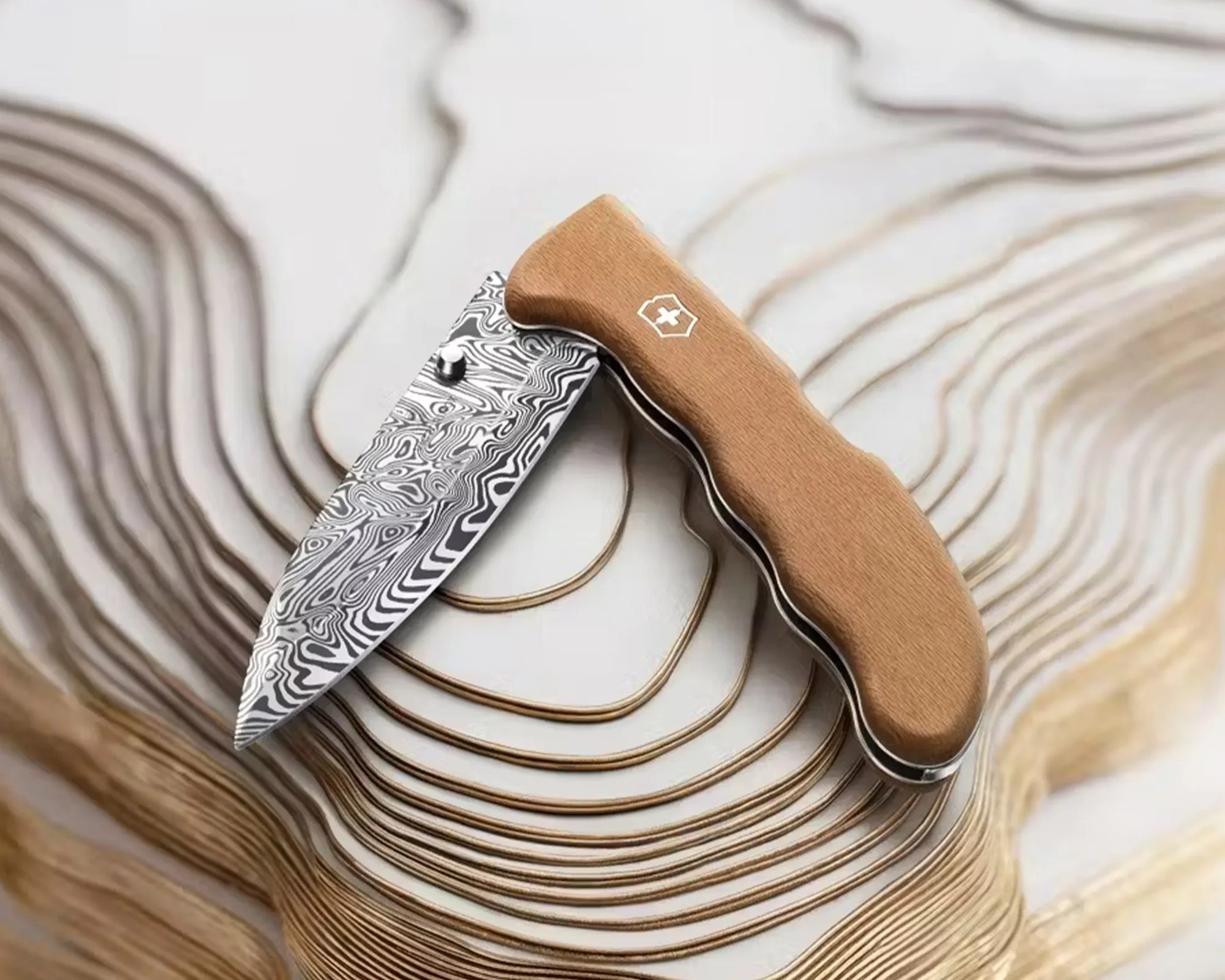 Folding knife with a patterned Damascus steel blade and a light brown wooden handle on a layered wood surface.