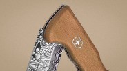 Close-up of a folding knife with a wood-textured handle featuring a silver cross emblem and a patterned black-and-white blade.