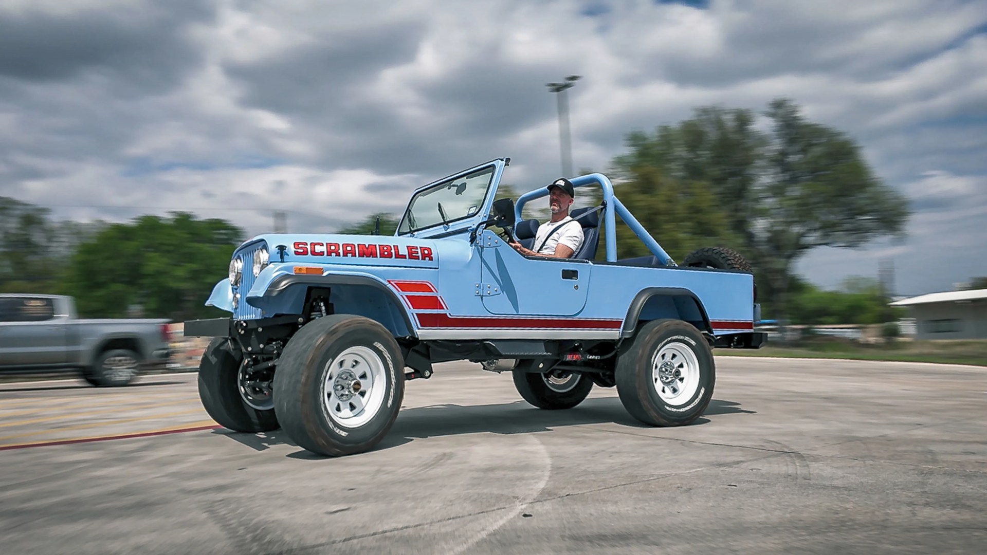 Light blue Jeep Scrambler with red stripes and large off-road tires driving on pavement.