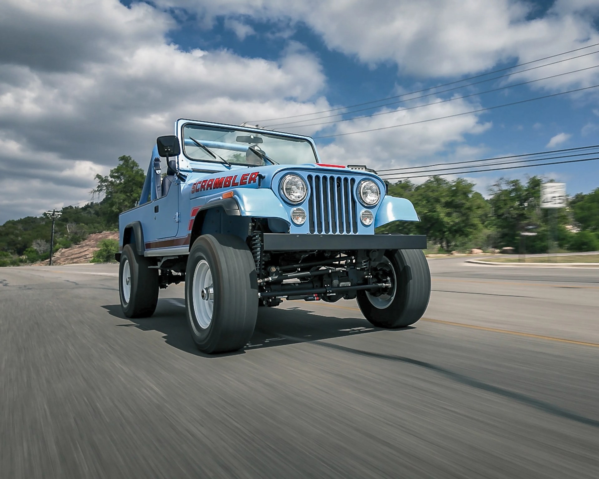 Light blue Jeep Scrambler with red lettering driving on a road under a partly cloudy sky.
