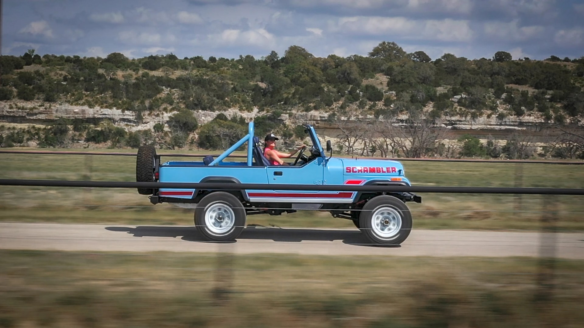 Blue Jeep Scrambler with red stripes and a person driving on a rural road with hills and trees in the background.