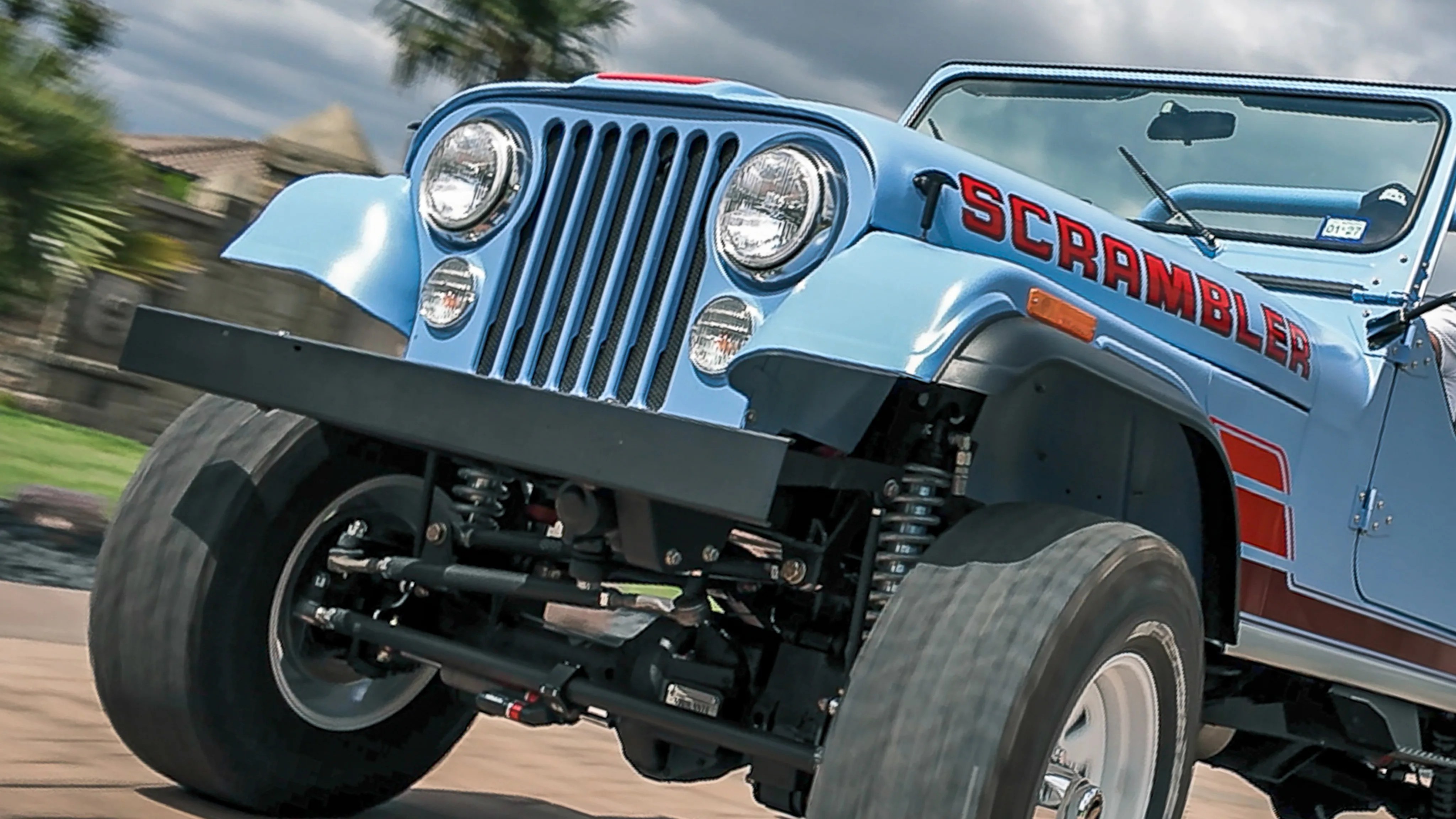 Light blue Jeep Scrambler with red lettering on the hood, large off-road tires, and black front bumper.