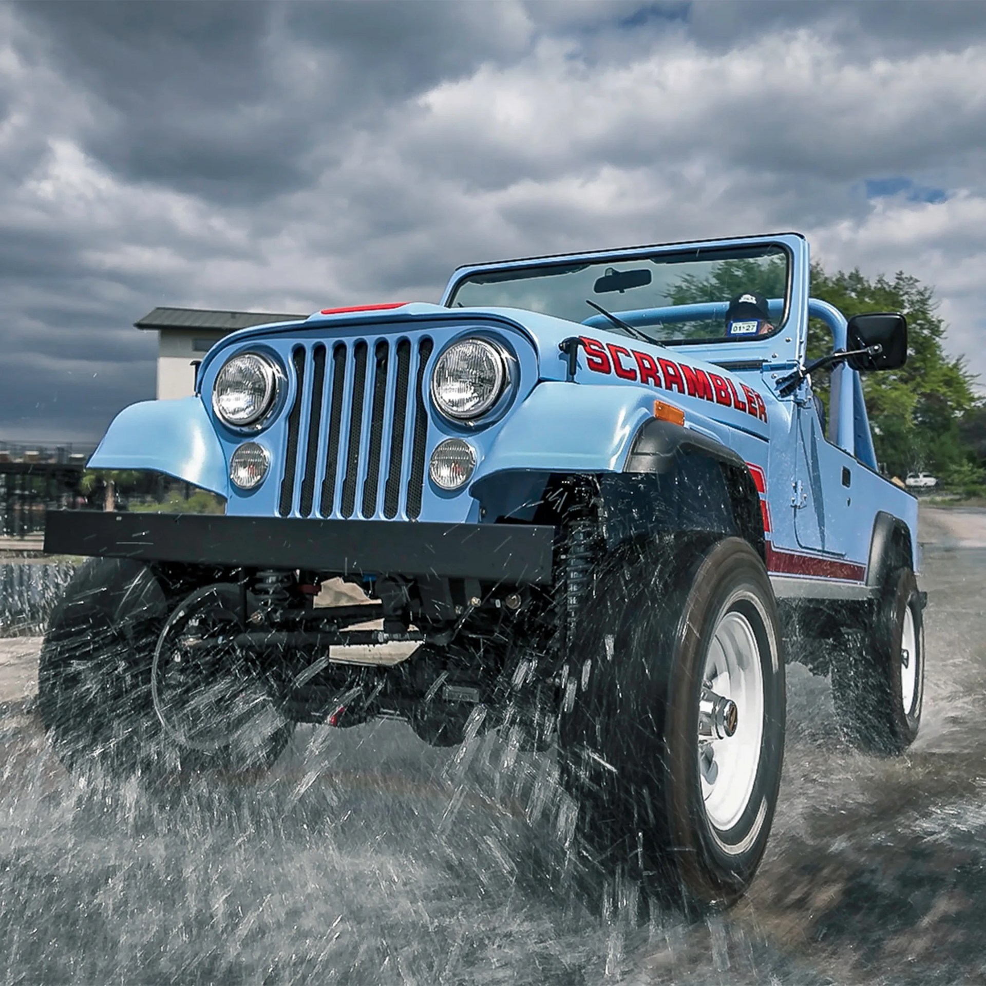 Light blue Jeep Scrambler driving through water splashing up around the tires under a cloudy sky.