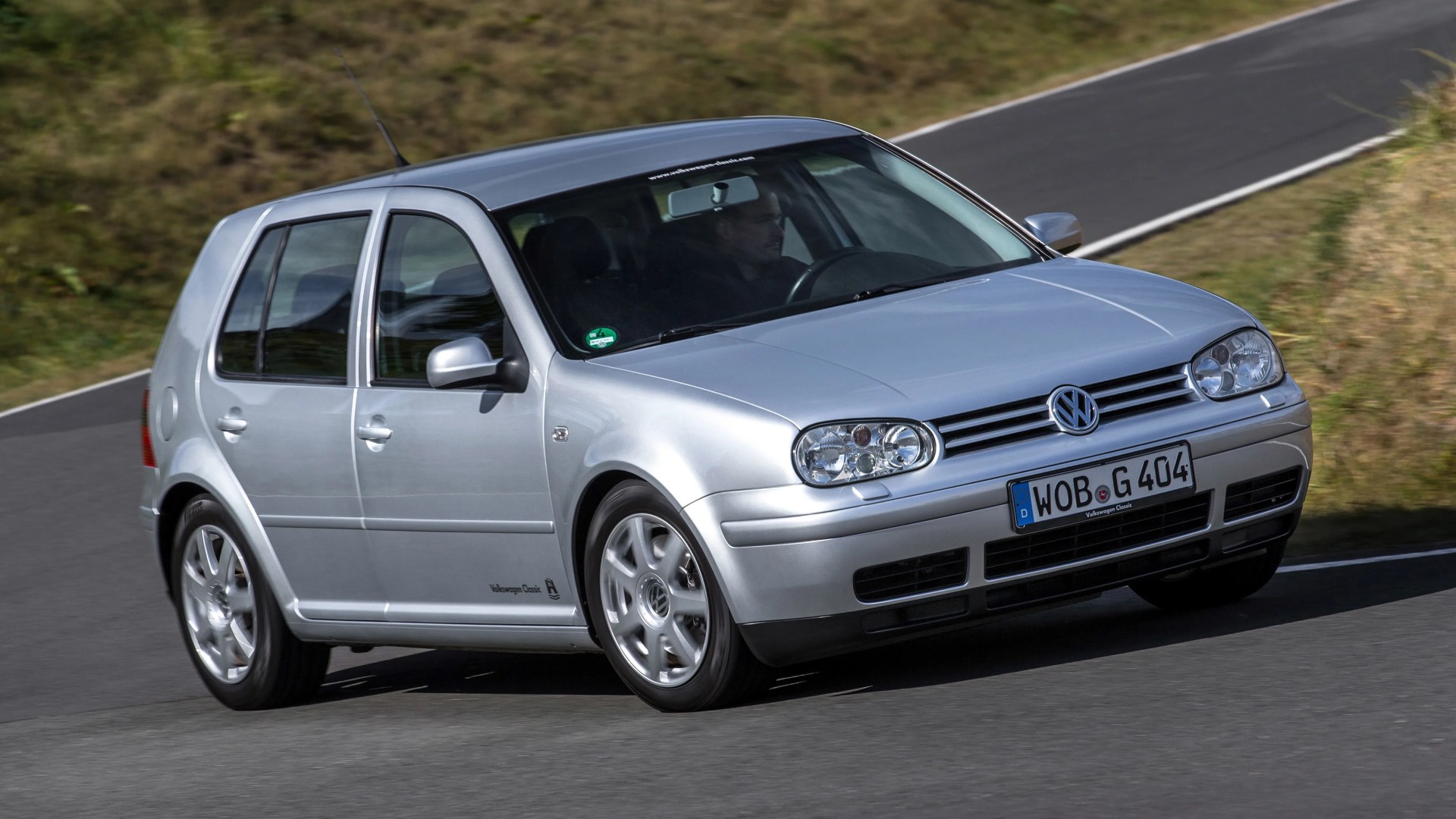 Silver Volkswagen Golf hatchback driving on a curved road with grassy surroundings.