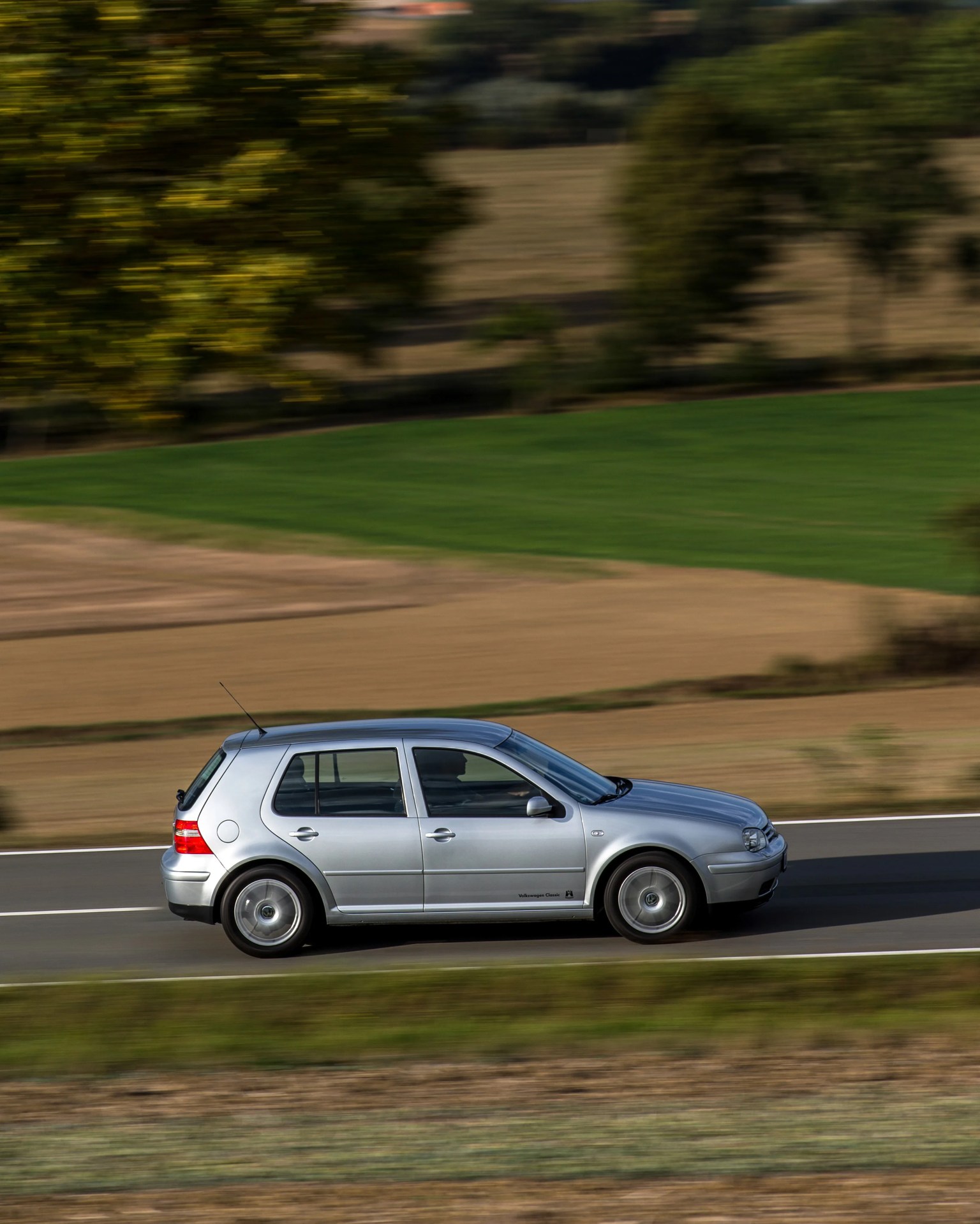 Silver Volkswagen Golf hatchback driving on a road with blurred green and brown fields in the background.