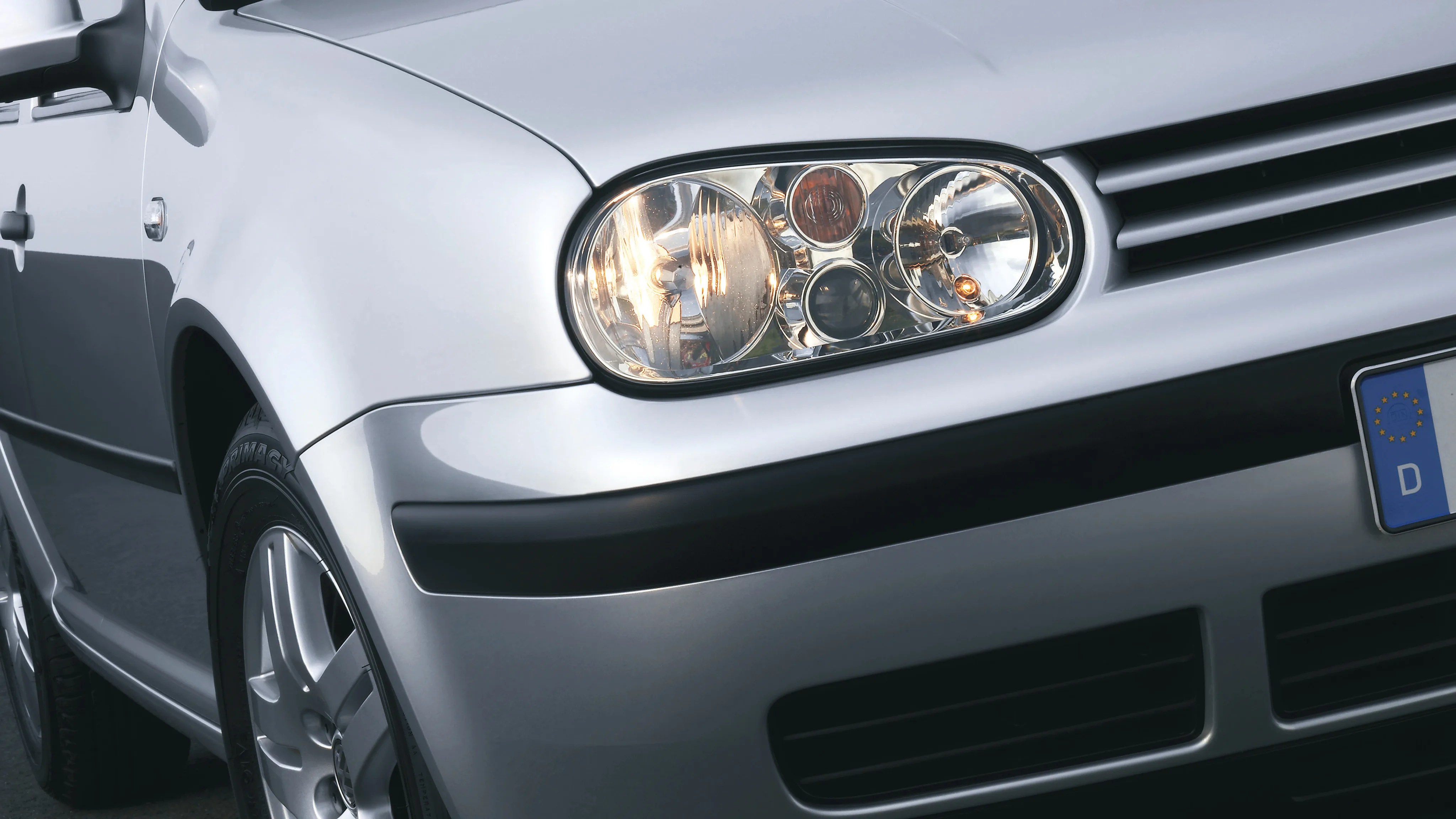 Close-up of a silver car's front left headlight, wheel, and part of the grille with a European license plate.
