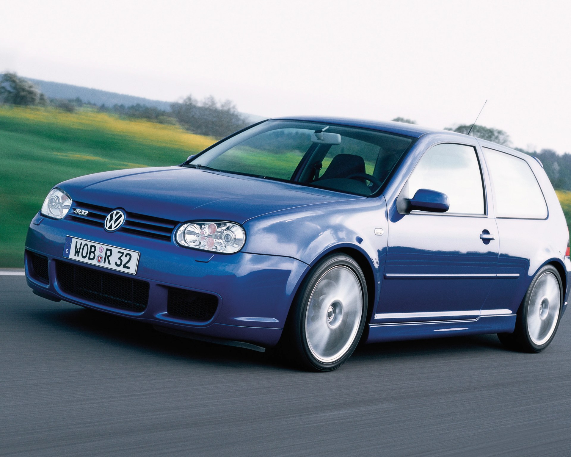 Blue Volkswagen Golf R32 hatchback driving on a road with blurred green and yellow landscape in the background.
