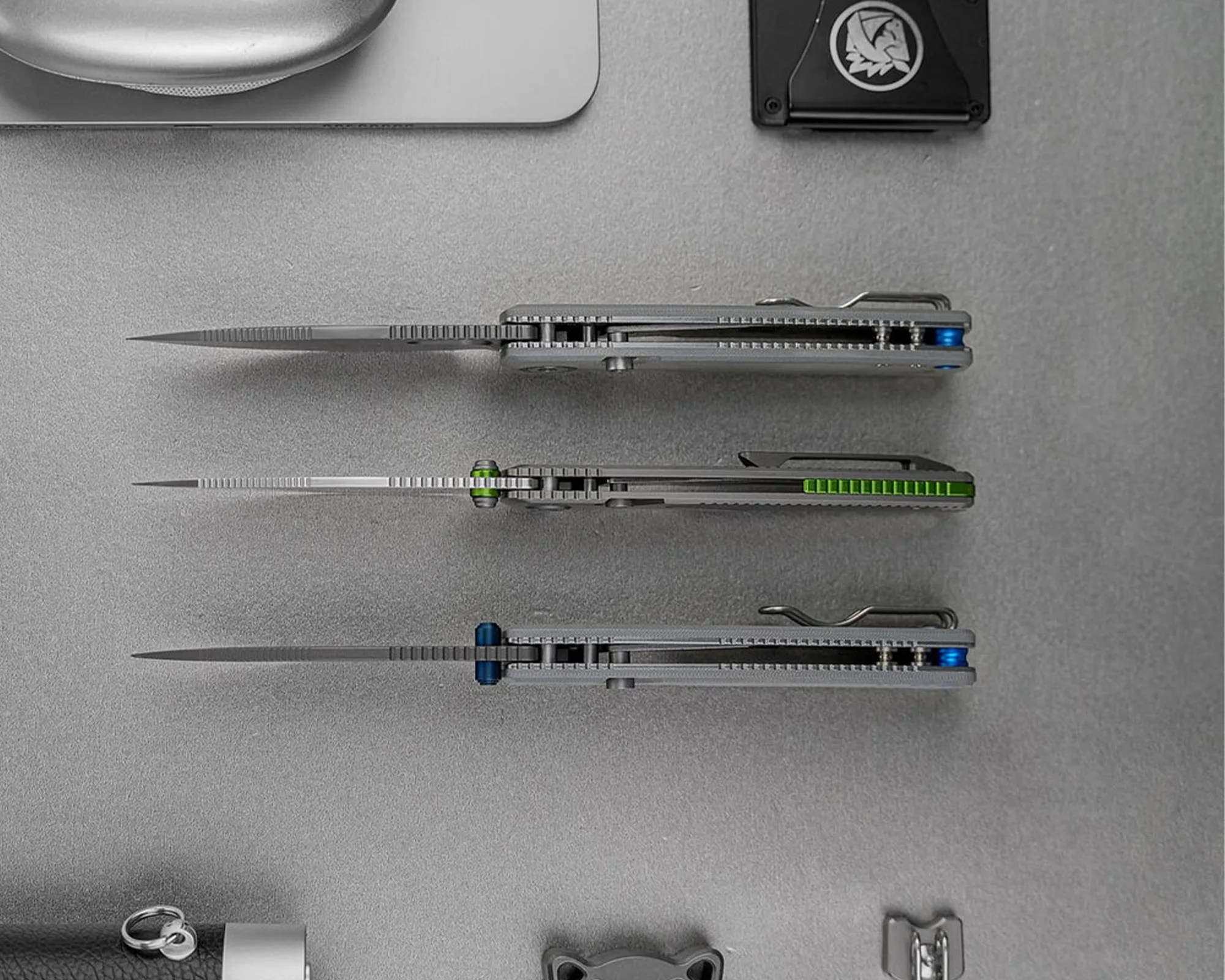 Three folding knives with partially serrated blades and colored thumb studs (blue, green, blue) laid flat on a gray surface.
