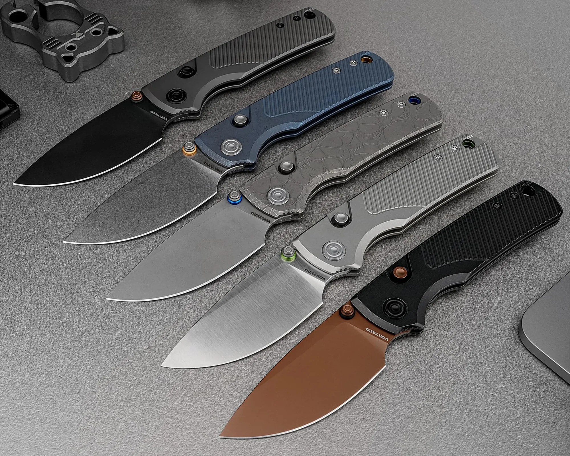Five folding knives with different blade finishes and textured handles arranged diagonally on a gray surface.